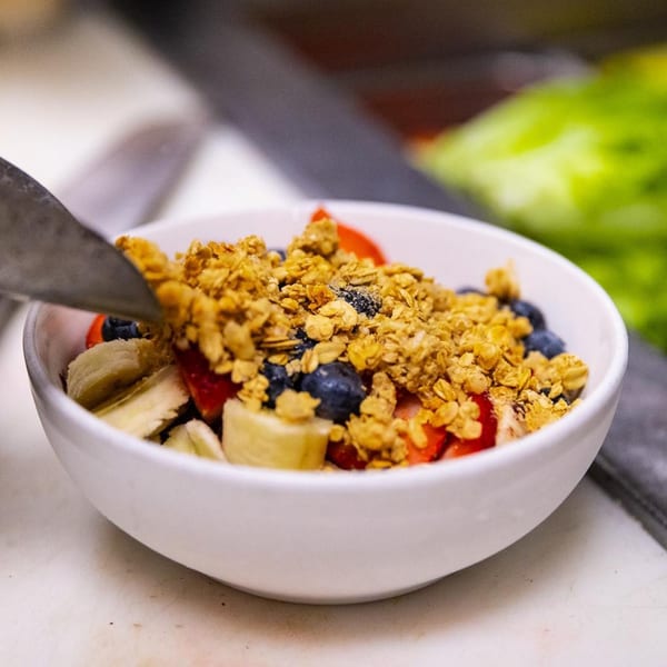 A white bowl filled with sliced bananas, strawberries, blueberries, and granola, with a spoon.