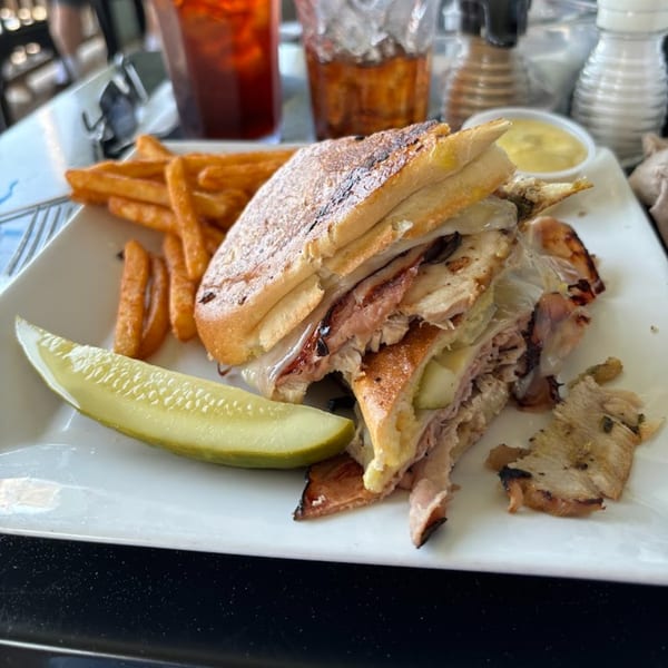 A Sunnyside Cafe sandwich with fries, a pickle spear, and dipping sauce on a white plate.
